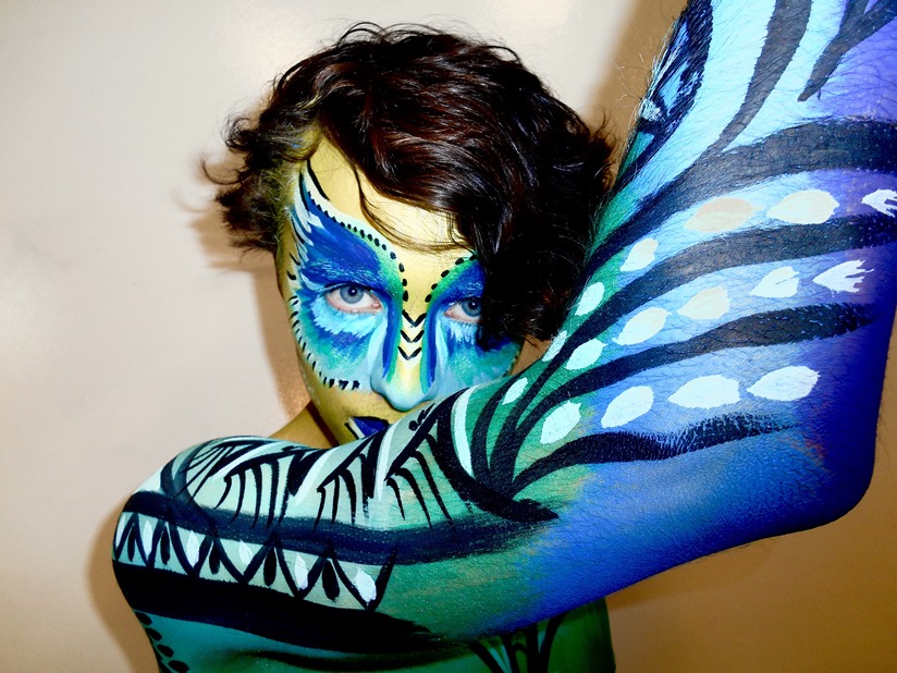 Close up of face and upper body of a person with body paint in blue, green, black and white and looks a bit like feathers.