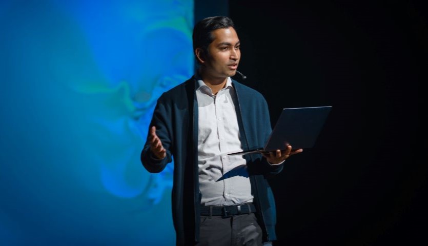 A man stands in front of a blue, projected backdrop, wearing a suit and holding a laptop open. He looks out in front of himself and holds with free hand as if speaking to an audience.