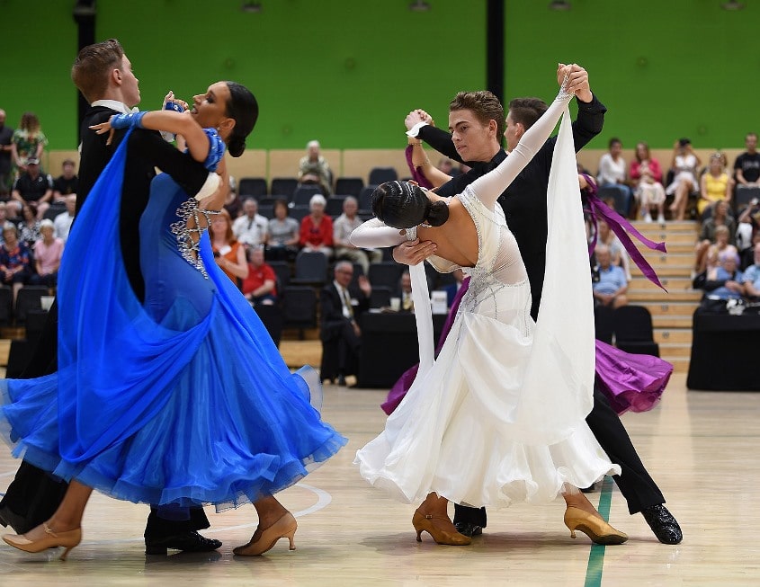 Two ballroom dance couples mid dance. One woman wearing is a white dress and the other is wearing a blue dress. Both male dance partners are wearing black.