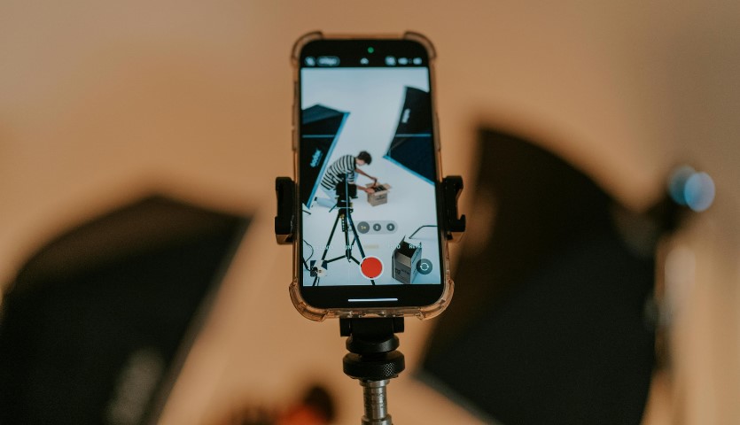 A smartphone sits on a tripod, showing an image it is currently recording of a young man bent over a box