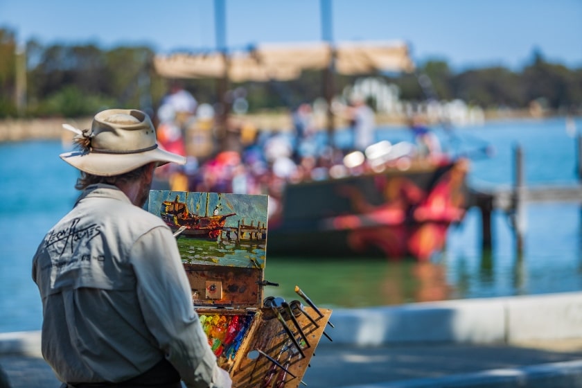 Male artist sitting by the water painting a pirate ship