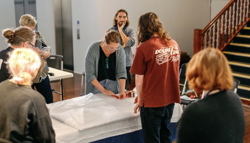 A group of people stand around a long white table, while one woman demonstrates unwrapping a framed piece of artwork
