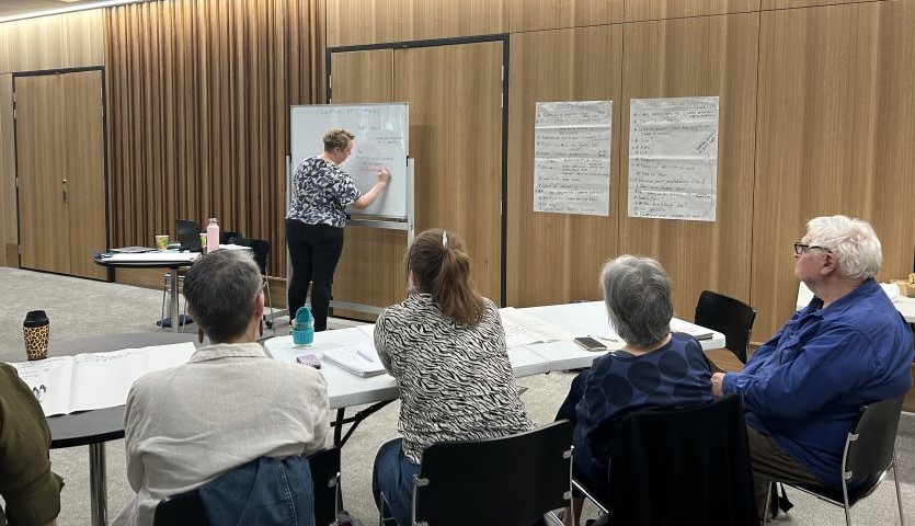 The backs of four people sitting at a table with paper in front of them, looking at one woman who is writing on a white board at the front of the room