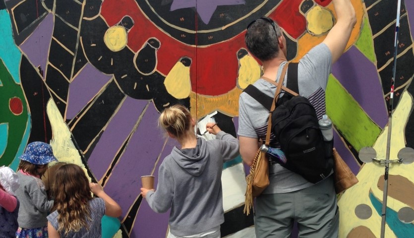 A man and three children are painting a wall using pots of colour, filling in shapes that have been pre-drawn.