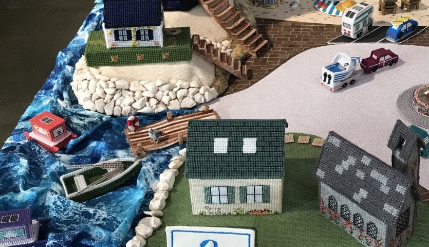 A close up picture of a miniature 3-dimensional coastal village scene, made of cross-stitched buildings, boats, cars and figures