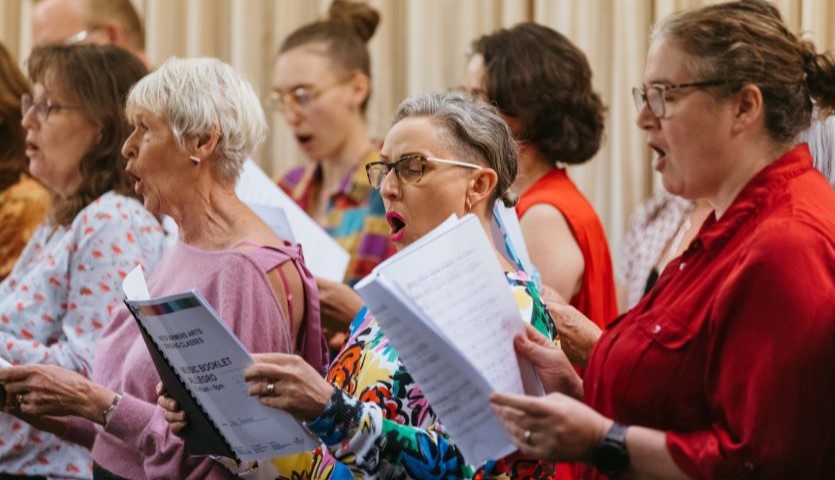 A group of people are standing together, all with their mouths open wide as though mid-song, looking down at open musical score books.