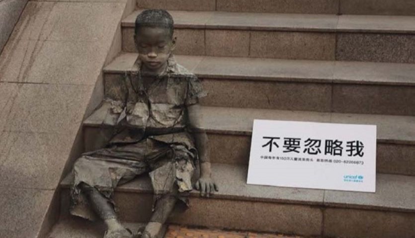 A boy sits on a set of stone steps, with a sign next to him showing Asian text. The boy is semi transparent so that you can see the steps through him.