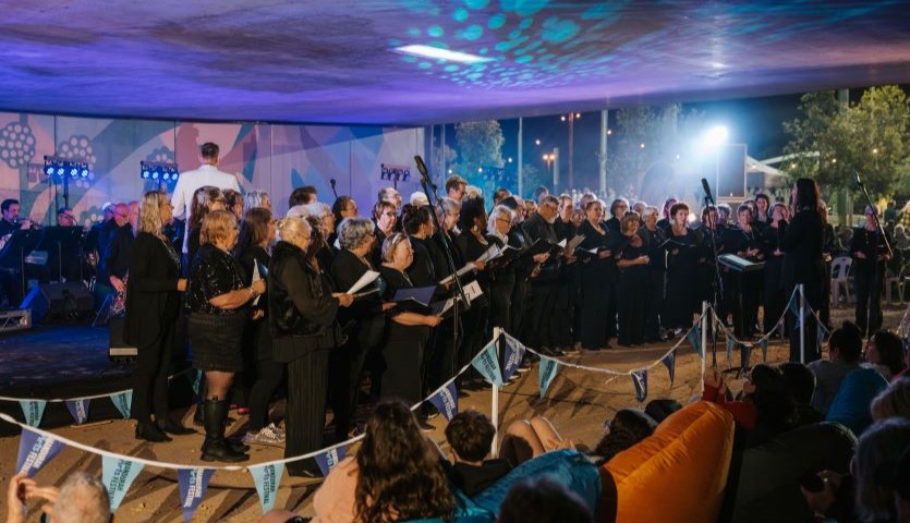 A group of men and women wearing black clothes stand in front of an audience, singing. They are surrounded by dramatic blue and purple lighting.