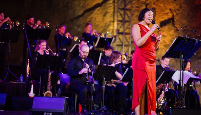 A band of jazz performers are lined up on a stage, playing their instruments while a woman in a red jumpsuit sings into a microphone.