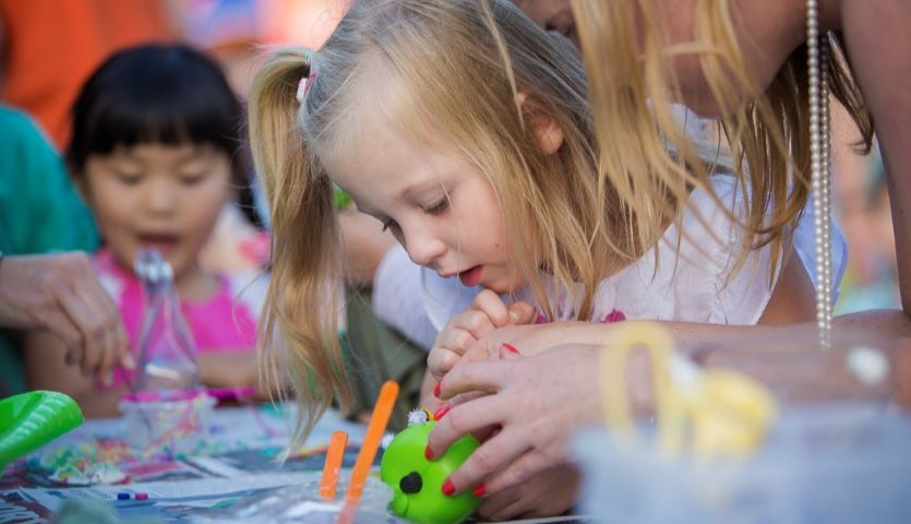 A small child is making a small green puppet with the help of an adult who is leaning over her. There are other children in the background.