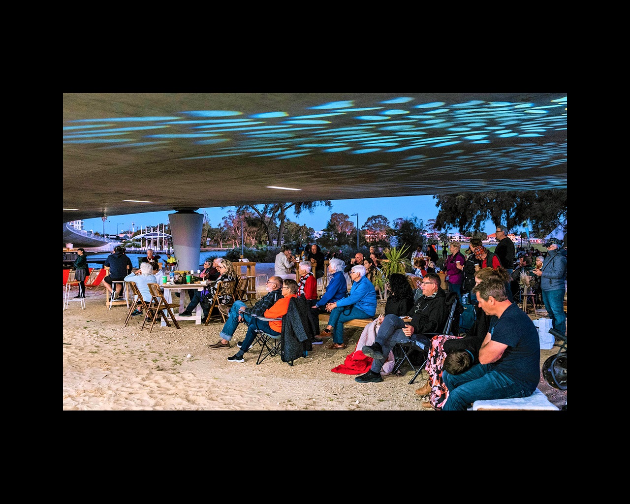 a crowd of people sit underneath a concrete bridge with theatrical stage lighting, watching a performance that is just out of frame