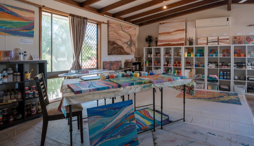 An artist studio with a large window, paint supplies in a bookcase along the wall and Aboriginal paintings on the table.