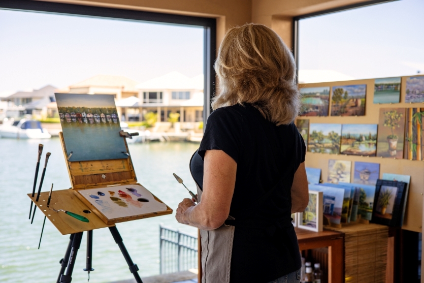 A woman with blond hair stands in her artist studio overlooking the water, with a painting of the scene that she is working on 