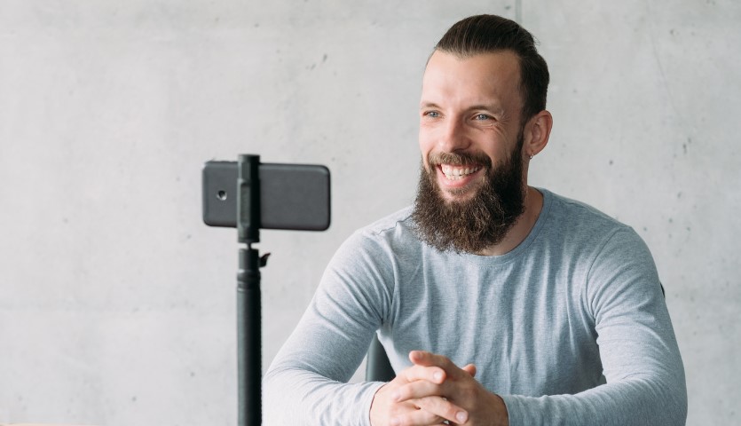 A bearded man in a grey shirt sits smiling in front of a smartphone on a tripod, as though about to record a video