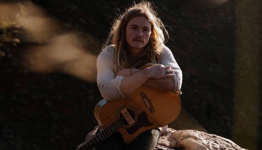 A young man sits in a lowly lit outdoor setting with a guitar on his knee