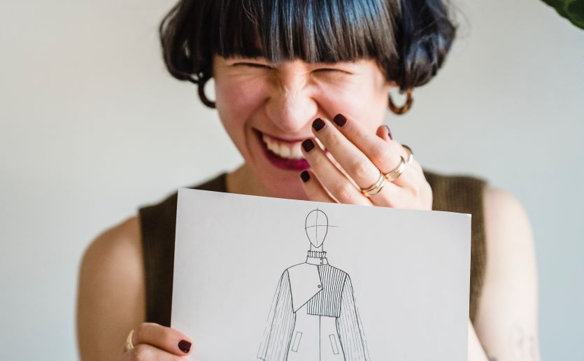 A woman with short dark hair giggles while holding up a sketch of a person 