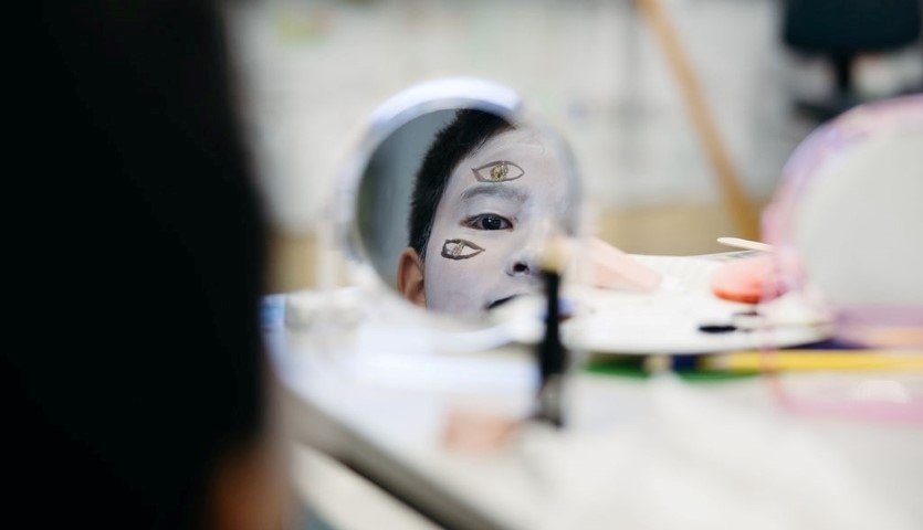 A small makeup mirror shows the reflection of a young person in white stage makeup, in the background a table is filled with makeup equipment