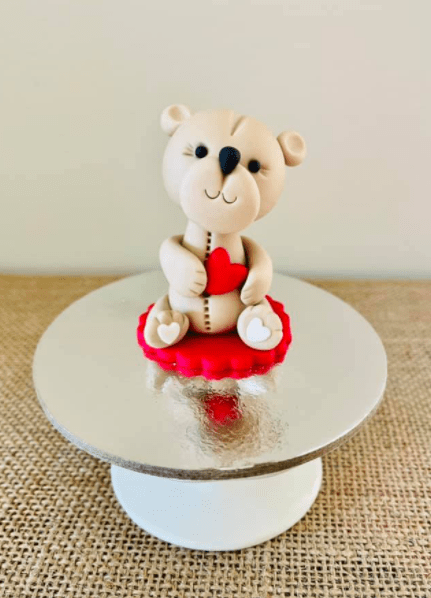 A white fondant teddy bear with a red love heart sitting on a silver pedestal.