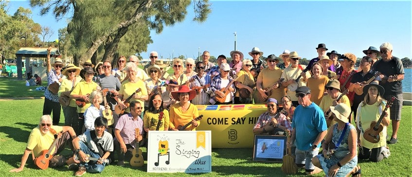 Large group of men and women primarily wearing yellow, gathered outside holding their ukuleles