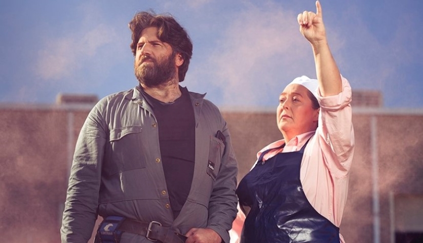A man wearing a blue jacket and a woman in a pink shirt and blue shiny apron stare into the distance. the woman is holding her hand in the air as though saluting something