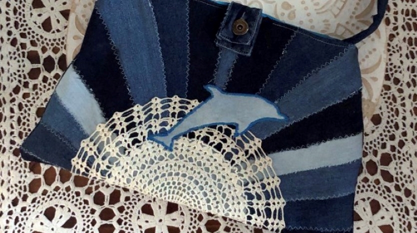 A bag made from several different blue denims with an applique dolphin lies on a table with a lace tablecloth