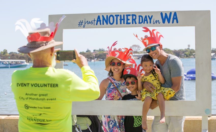 A woman speaks to two event volunteers wearing bright yellow shirts with 'City of Mandurah' printed on the back, inside a storage or event setup area.