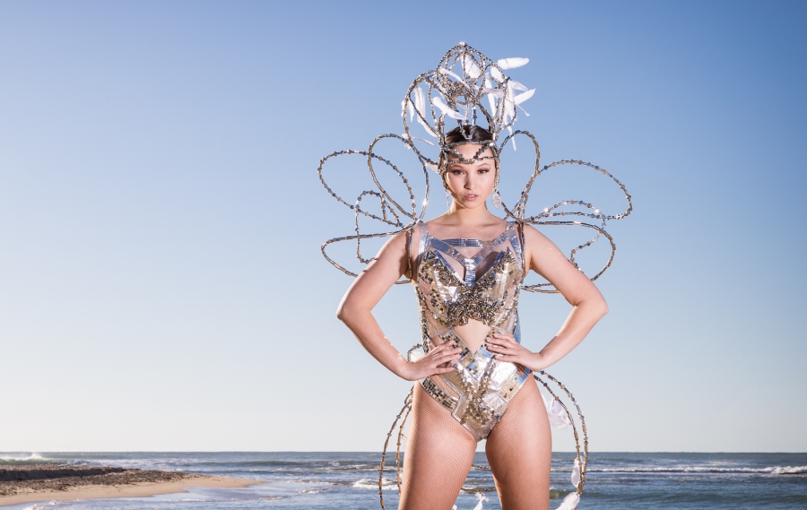 Wearable Art Mandurah 2021 garment called Light by Janelle DSouza. The model is wearing a silver garment with detailed design and headpiece. Photo taken at beach with clear, blue sky.