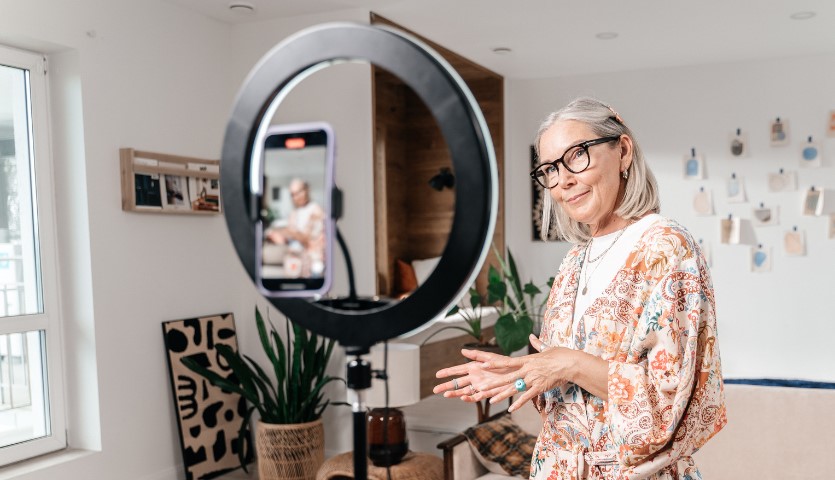 A woman stands in front of a light and a smartphone that is filming her, hands outstretched as if demonstrating something
