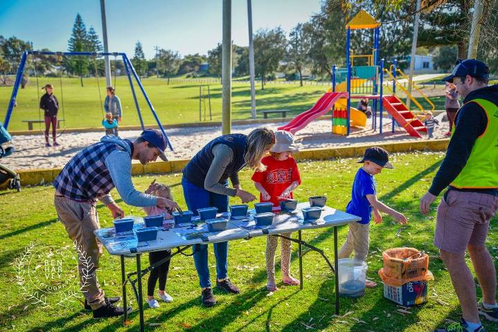 Adults and children doing activities on a table outside, with a children's playground in the background.