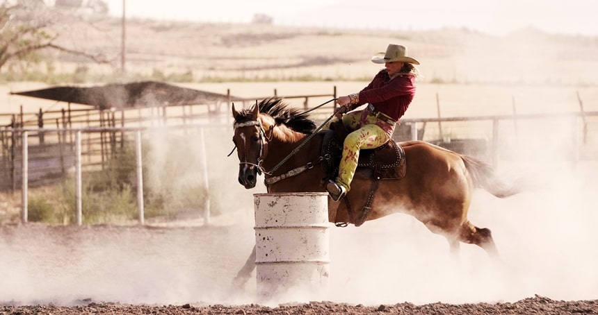 A rodeo woman on horseback barrel racing around one white barrel with lots of dust in the air.