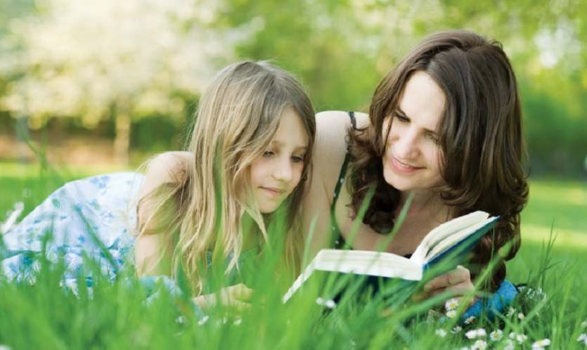 A woman and young girl lying on their stomachs in the grass reading a book together.