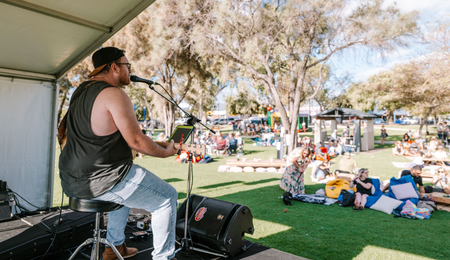 Musician playing guitar onstage in front of a small crowd on a sunny day