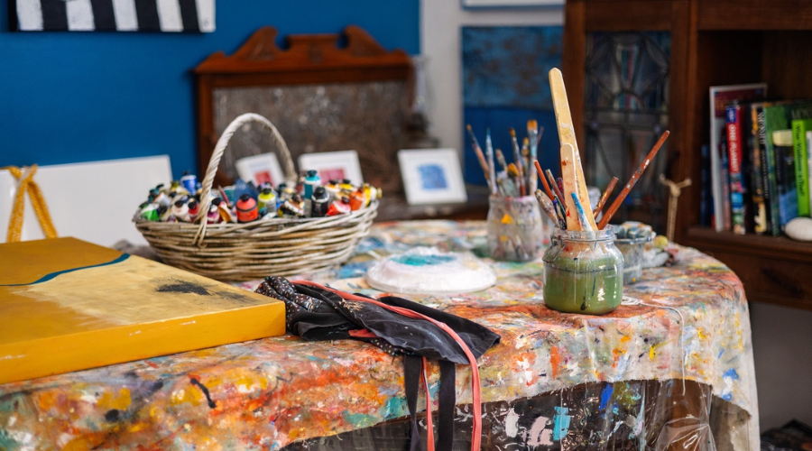 Landscape image of art supplies including paints, brushes, aprons and canvas scattered across a desk