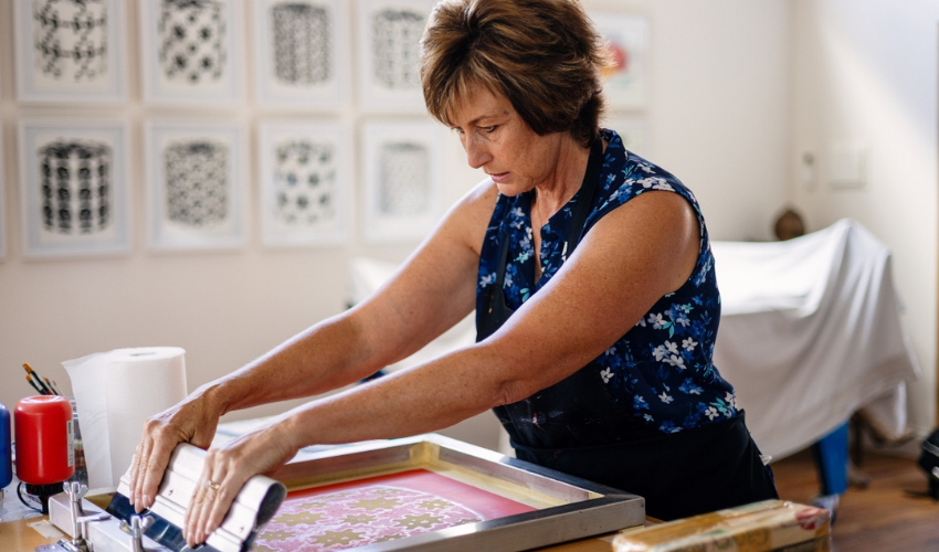 A lady standing at a waist height table as she creates an artwork using paint and a printing press