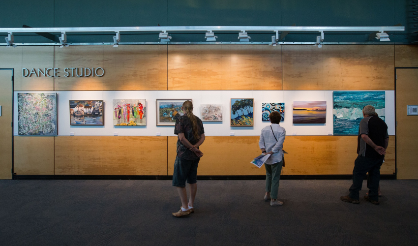 Three individuals stood inspecting a line of artwork hung on the wall directly before them