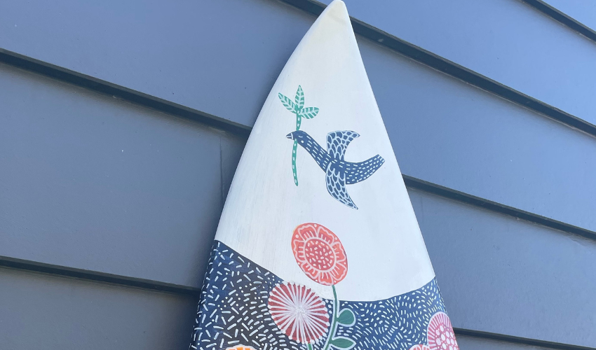 Image showing the top half of a blue surfboard illustrated with a bird and patterns leaning up against a slatted wall