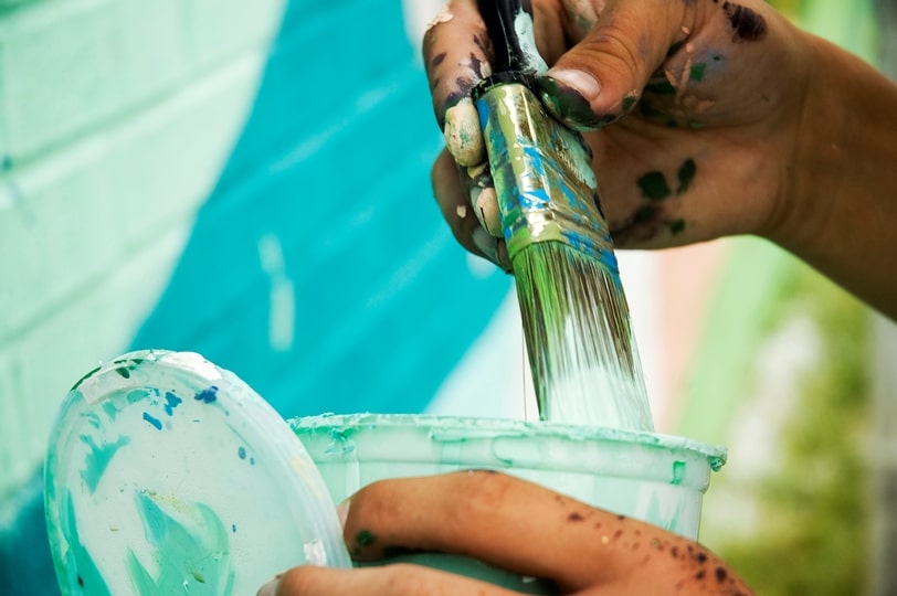 Close-up of a paintbrush with white paint being wiped on the side of a paint can or pot.