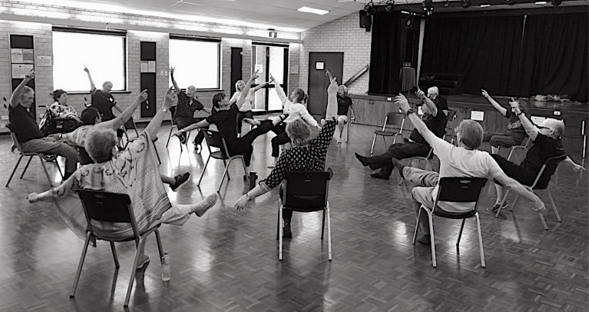 Black and white image of people dancing in chairs, set up in a circular pattern around the dance teacher