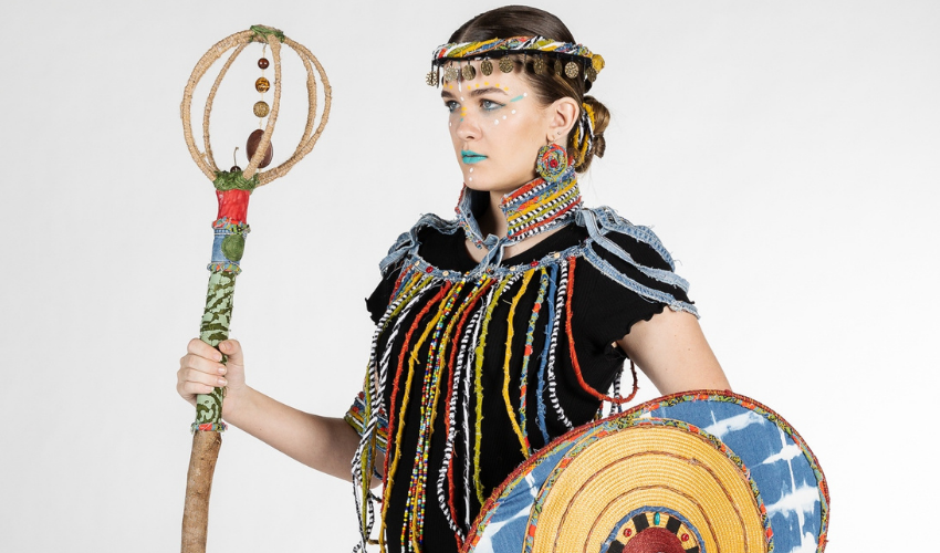 Close-up of a young female model from the waist up wearing a colourful decorative garment over her clothes and holding an ornamental staff
