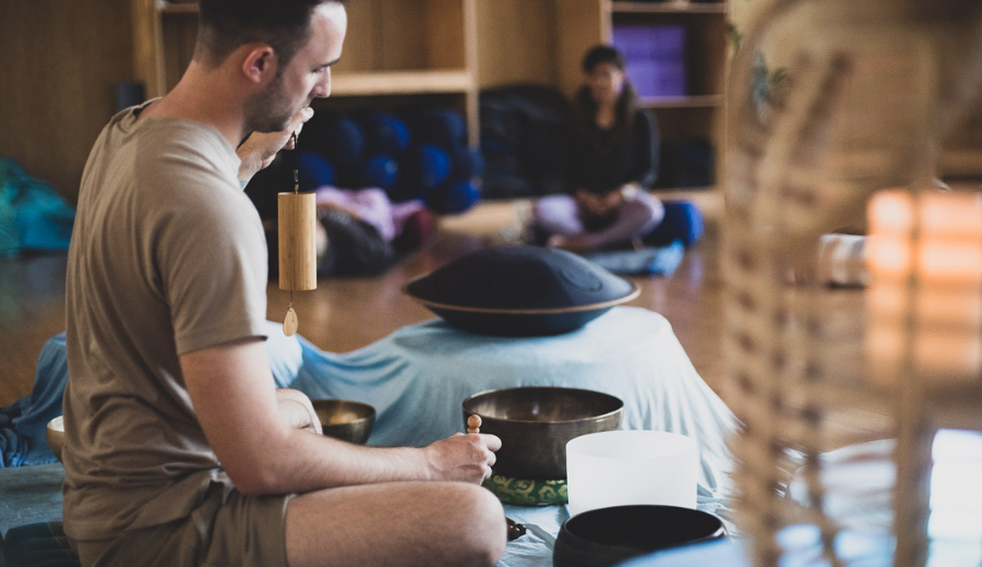 A young male is pictured in side profile, as he sits with legs crossed on the ground surrounded by yoga equipment