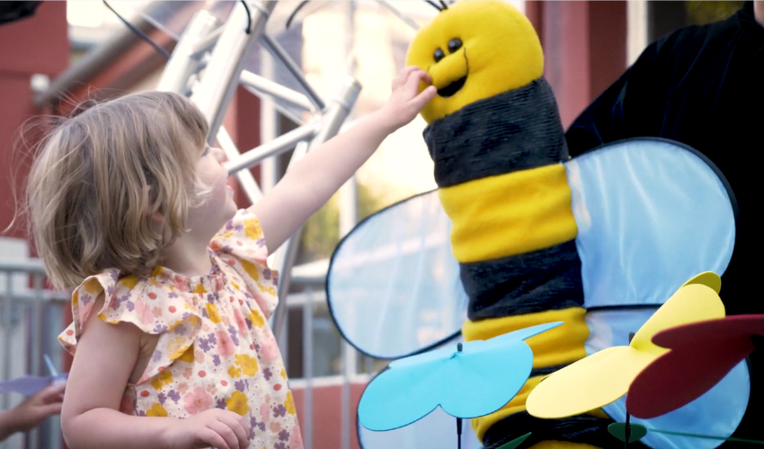 A young child reaching up to touch a tall, black and yellow puppet of a bee with wings