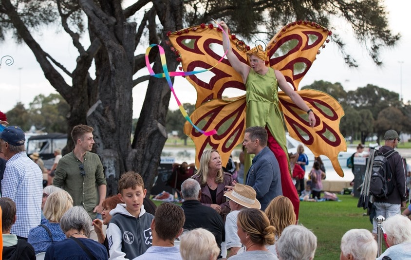 Stilt walker with large orange butterfly wings twirls a ribbon at people picnicking on the Eastern Foreshore