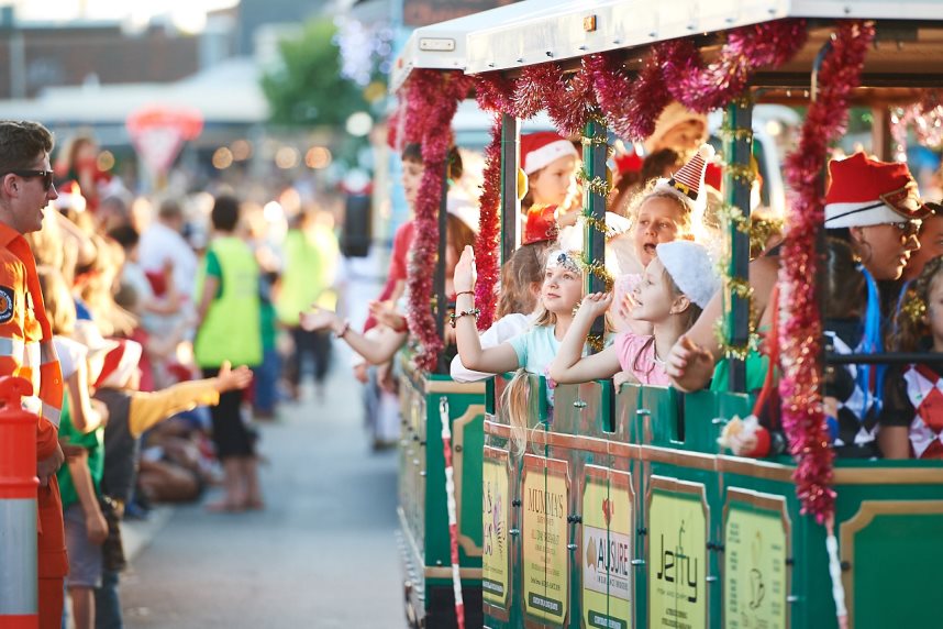 Children wearing Santa hats and tinsel sitting in the carriage of a fun train, waving at the crowd as it drives along the road.
