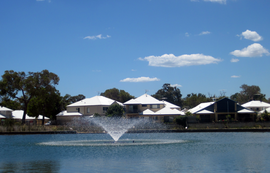 A fountain spraying water in the middle of a lake which is in front of suburban houses