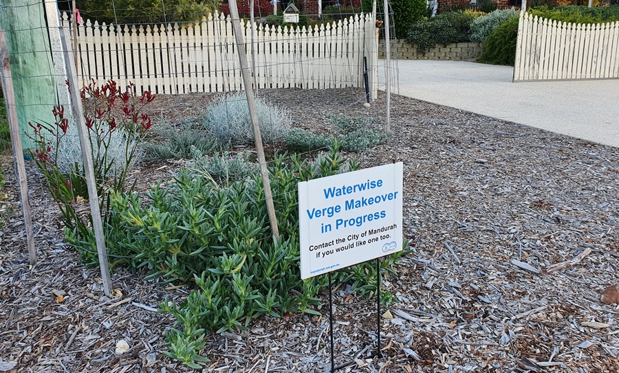 Sign that says waterwise verge in progress in front of a newly planted garden of waterwise plants protected by garden fencing on a large verge.