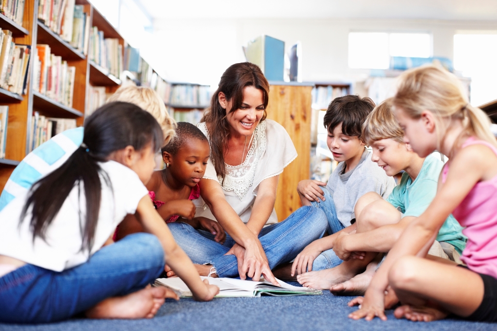 White woman with brown hair sitting cross-legged on a library floor, pointing at an open book, while 6 children of various races and colour block clothing also sit around her, looking