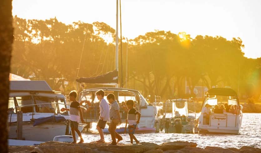 Four small recreation boats in the water during sunset while a group of young boys play on the shore.