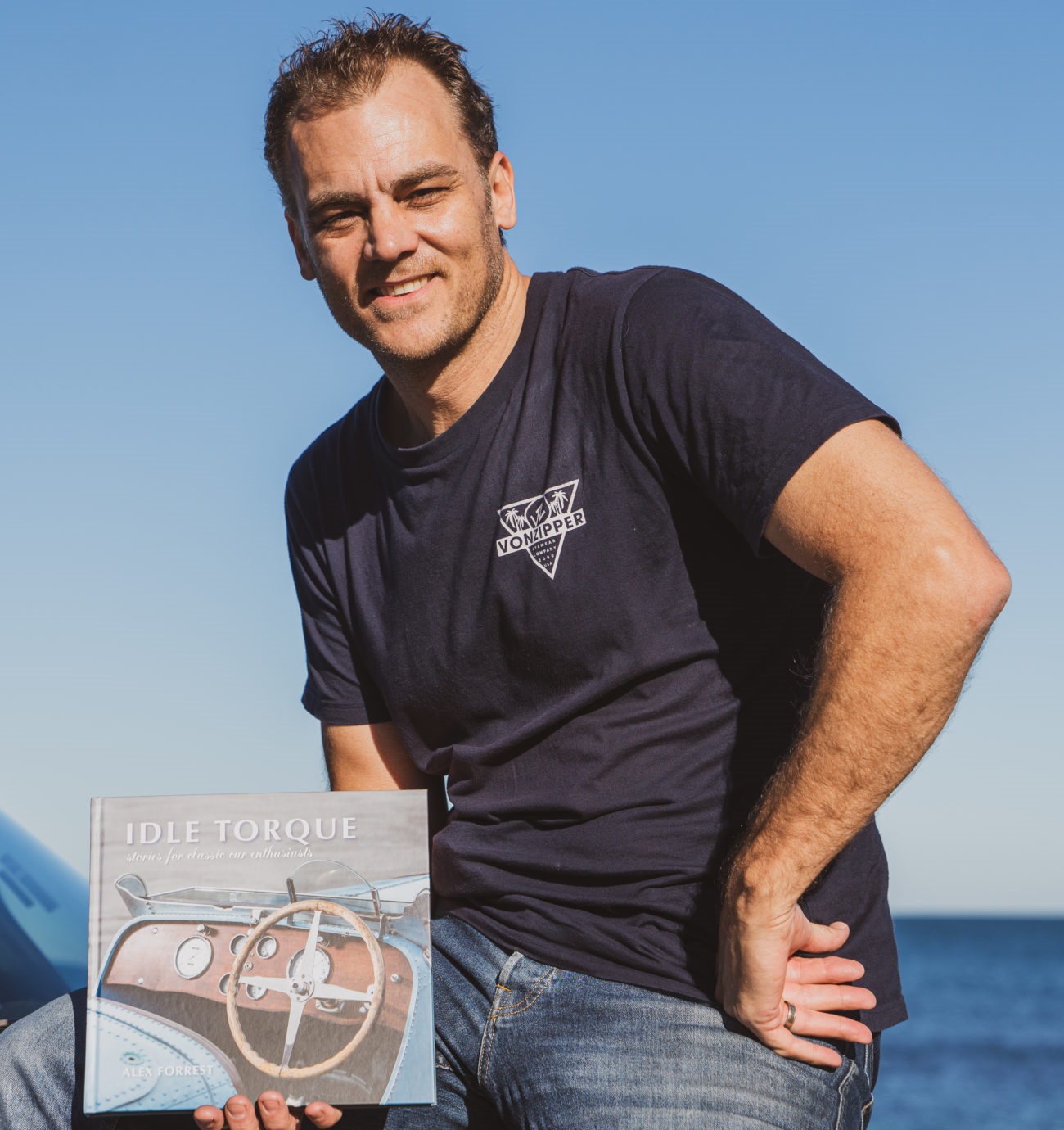 Author Alex Forrest wearing blue jeans and blue t-shirt holding a copy of his book Idle Torque