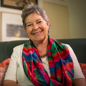 Author Sue Boyd wearing a white shirt and colourful scarf sat on a couch