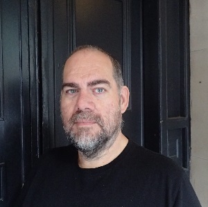 Close up head-shot of author Tim Richards in a black t-shirt stood in front of a black door